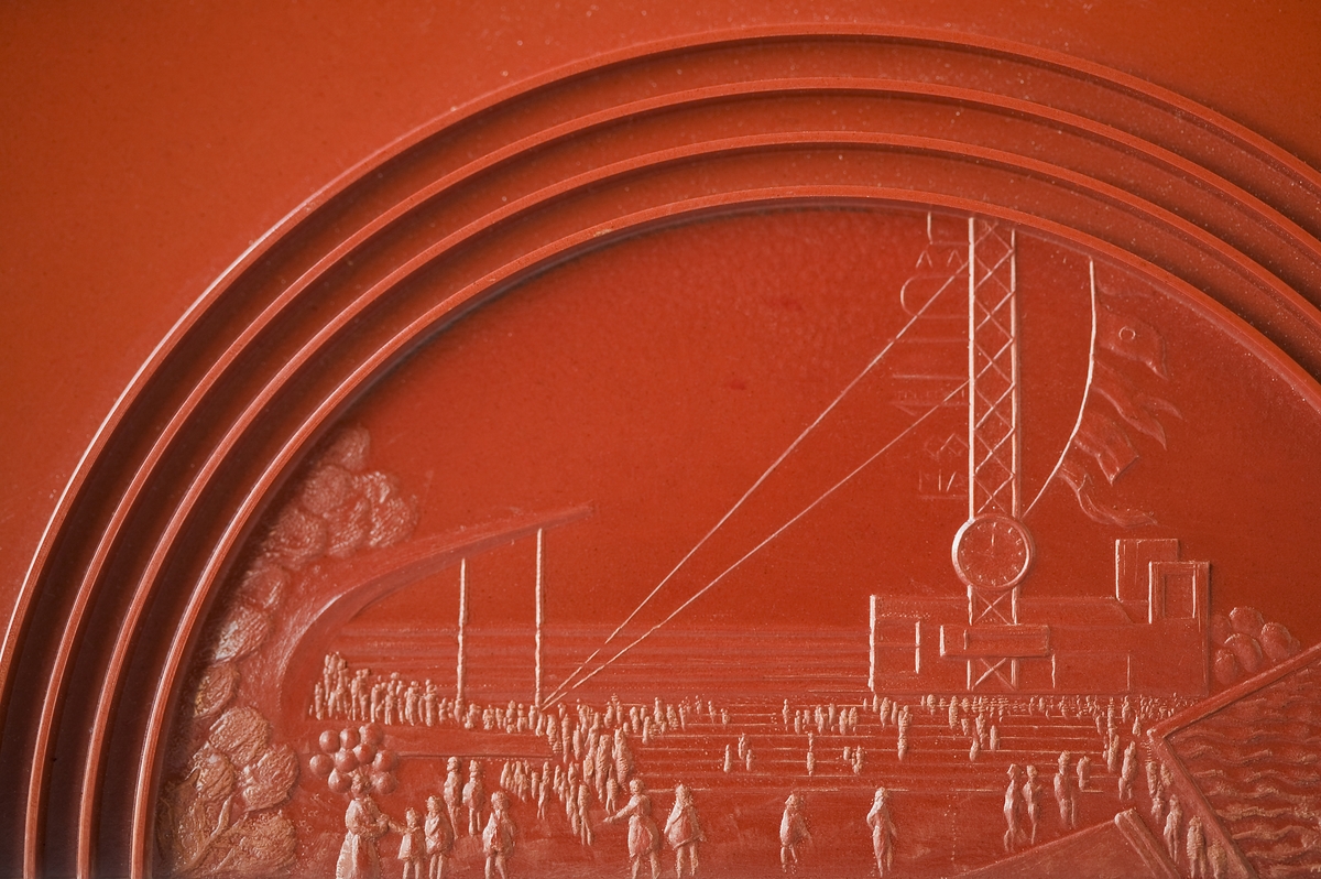 Cigarrskrin, pressat av röd pollopas som är en karbamidplast. Ytorna har upphöjda ränder som dekor. Locket med graverad reliefbild av Stockholmsutställningen 1930. Skrinet är signerat i botten.