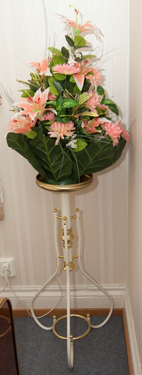 Pidestall med plastblomster i vase; hvit med rundt messingfarget fat i metallblikk.