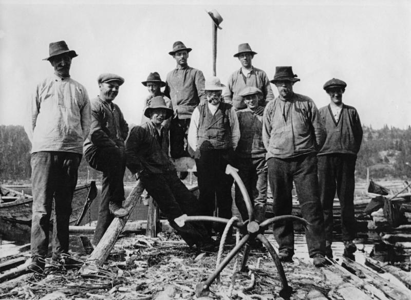 Fotografiet viser ti menn på en tømmerflåte, hvor det ligger et digert anker foran karene. Bak dem synes det å være bygd et stativ med en stolpe, hvor noen  har tredd en hatt på den høyeste stolpen.