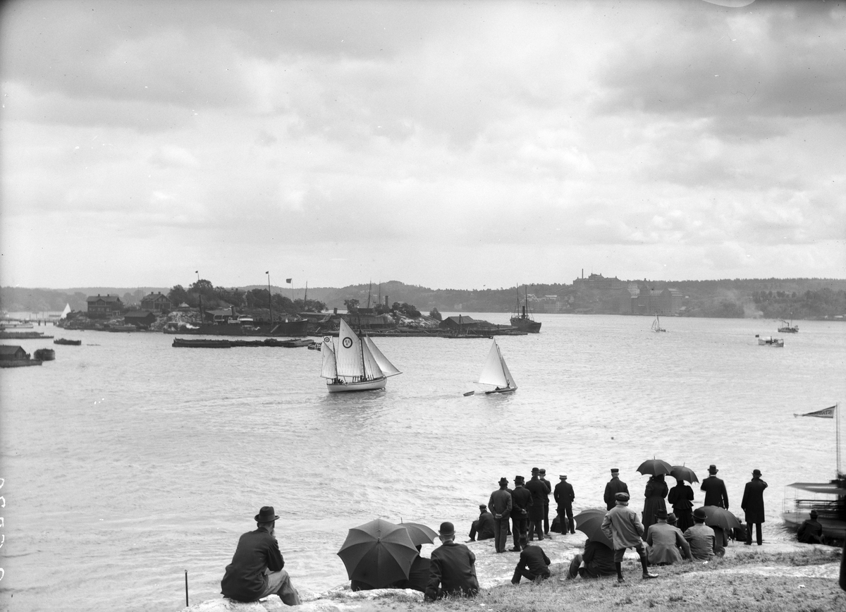 Vy från Kastellholmen mot Beckholmen med Saltsjö kvarn i bakgrunden. Sällskap på stranden. Omkring 1897.