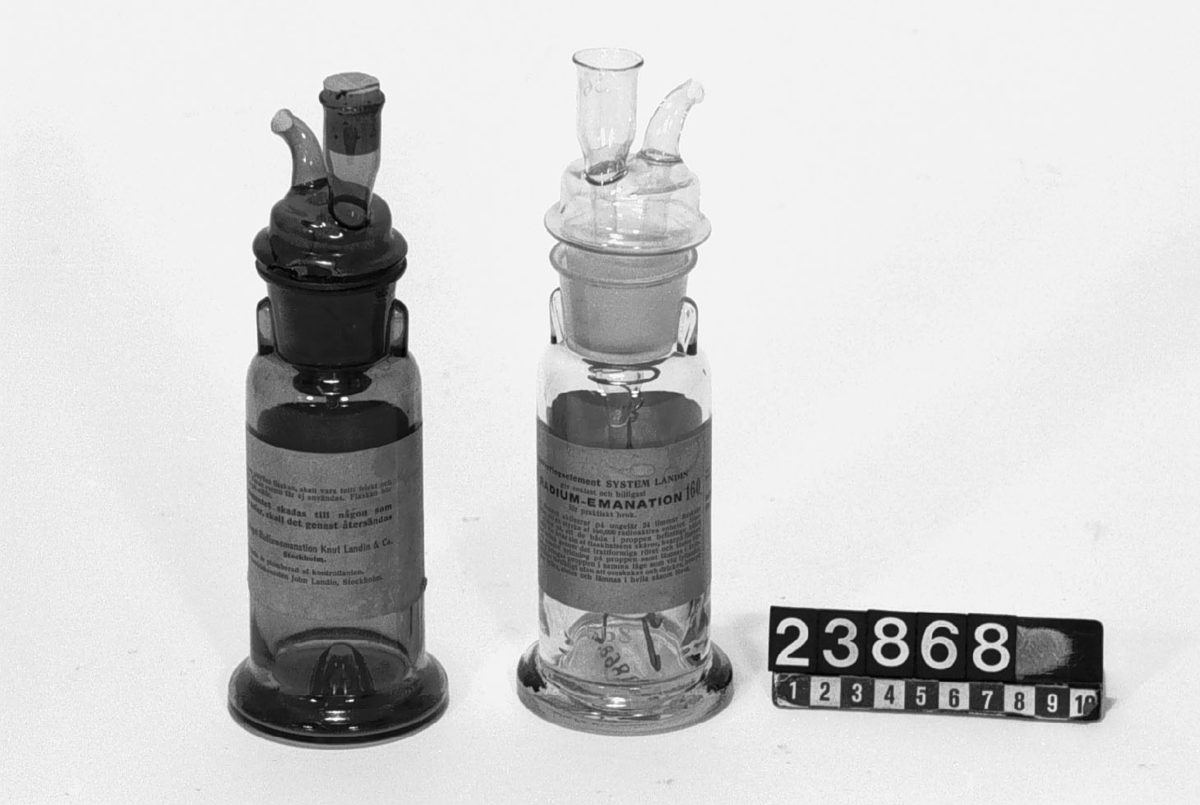 Två flaskor av glas med "aktiveringselement system Landin" för beredning av radioaktivt dricksvatten glasvis. Volym ca. 175 cm3. Propparna ha kanaler som medger hela flaskans fyllning med vatten samt nedtill fäste för hållare (av nysilver, TM 23.867) till det radioaktiva preparatet. Flaskorna är numrerade bl.a. för att ej förväxla propparna. Nr. 210 av brunt glas och är laddad med preparat med normaldos 10 000 radioaktiva enheters (voltenheters) styrka. Nr. 668 är laddad med 160 000 enheters styrka.
