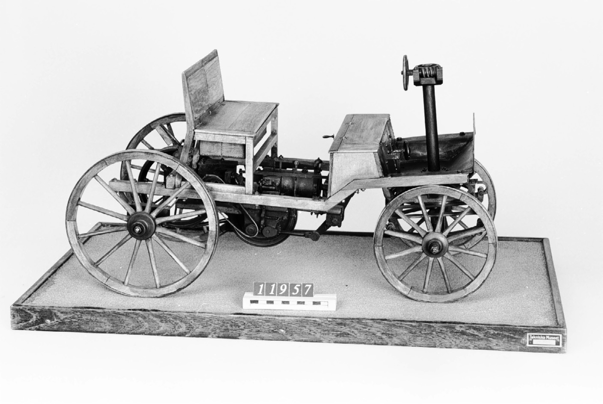 Modell i skala 1:5 av bensinmotordriven bil med hästvagnsliknande kaross. Motorn tillverkad av Märky, Bromovsky & Schultz 1888. Kraftöverföring från motorn med remmar till remskiva på bakaxeln. Ram av trä, framvagn med halvelliptiska fjädrar. Trähjul skodda med järnband. Förebilden har 2 m axelavstånd. Monterad på en platta 700 x 400 mm  Encylindrig luftkyld fyrtaktsmotor Effekt cirka 4 hk
