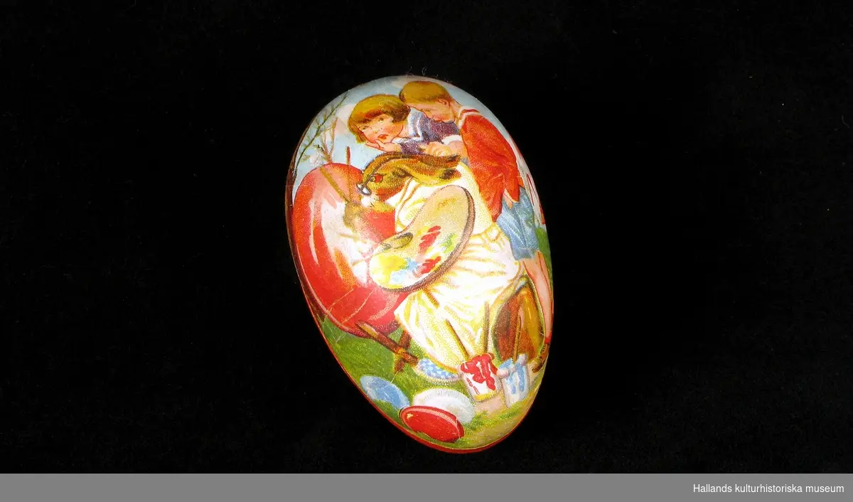 Påskägg av pressad papp i två halvor. Två mångfärgade bilder, på ena halvan drar två kycklingar en kärra med ett ägg på, hönan och tuppen ser på. Den andra halvan visar en hare som, utklädd till konstnär, målar ett ägg, två barn ser på.