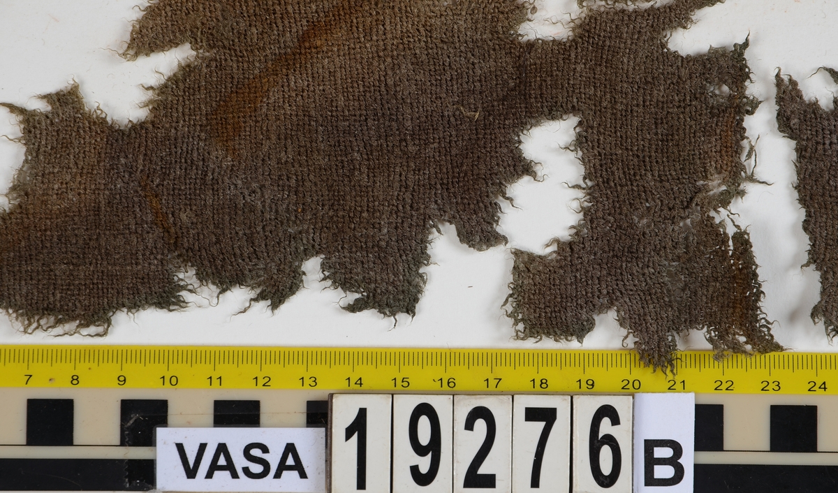 Fnr 19276a: Fragment av en tygbörs av sammet i form av en pung.
Fnr 19276b: Två fragment av ull vävt i tuskaft.

Textildokumentation 2017:
Fnr 19276a: Består av tre hopsydda delar (160 mm, 50 mm och 49 mm). I nuläget 140 mm lång. I överkant tydliga avtryck av veckning. Troligen har ett avvikande tyg varit fastsytt upptill, märken efter stygn finns på några ställen. Vid överkanten på den större delen är det inskarvat en trekantig bit av samma tyg. 

De båda mindre bitarna har stadkanter, dessa är i spetskypert och inslagstrådarna sticker ut på sidorna. 

Delarna är hopsydda med kaststygn från avigsidan. Sömsmånerna är nedsydda med förstygn. Mellan den stora delen och den mindre är det bitvis sytt med efterstygn. Sytt med en tvåtrådig tråd av silke, för blotta ögat ser tråden svart ut, i mikroskop ser den blå ut. 

Förstygnssömmarna fortsätter nedanför sammeten mellan de mindre delarna. Längden på pungen kan inte ha varit mindre än 165 mm plus en överkant med dragsko. 

Sammeten är skuren och oskuren, troligen dessutom i olika höjd. Samtliga delar har luggen riktad nedåt. På den lilla skarvbiten är luggen riktad lite åt sidan. 

Fnr 19276b: Två fragment. Det större fragmentet har kant tillskuren på skrå.