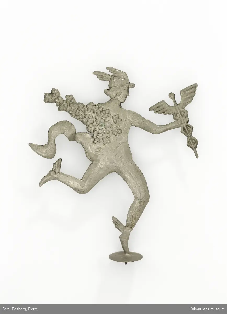 KLM 28228:9. Prydnadsfigur, till blomsterdekoration. Av metall, tenn. Figur föreställande en man, guden Hermes i grekisk mytologi. Han har hjälm och skor med vingar på och håller i en spira med vingar. Hans bål och ena arm är inlindande i blommor. Hans kläder rör sig i vinden. Han står på ett ben, benet står på en rund platta med skruv och mutter på undersidan.