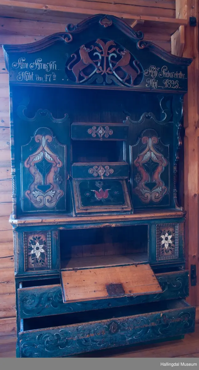 Delene satt sammen v.hj.a. tapping og trenagler.  Gavlen buede sider med skåret kronlist, delt i 3 profilerte, med opprullede ender.  Kronlister fortsetter på sidestykkene.  I toppen av kronlist hjerteformet, gj.brutt utskjæring opp ned.  Gavlens sentralfelt utskjæring i dyp flatskurd.  To løver, vendt mot hverandre, griper om hellebordskaft.  En stake som øverst kløyves i to bjørnehoder i profil skiller løvene.  Gavlen (el. baldakinens) forplate danner fremsiden i skuff i skapets bredde og dybde.  Overskapet:  Åpen hylle øverst, ca. 25 cm høy.  Under hyllen er det delt i 3 felt.  Ett skap i hvert sidefelt.  Midtfeltet har 2 skuffer adskilt av åpen hylle.  H. Skuffe: 12,5 cm.  H. åpent rom: 17 cm.  Under nederste skuff, skap med skråsider som stikker ca. 15 cm ut fra forveggen på overdelen.  Skapet dekkes av liggende skråklaff.  Festet jrenbeslag og hengsler nederst.  
En stor skuff i hele skapets bredde, 6 cm høy nederst, 32 cm dyp.  Over skuffen er rommet delt i 3 felt.  Hvert sidefelt har 2 skuffer og midfeltet en dyp skuff.  Midskuff har fylling med utskåret medaljong, klaff og hylle og skuffen rødmalt. .................................................