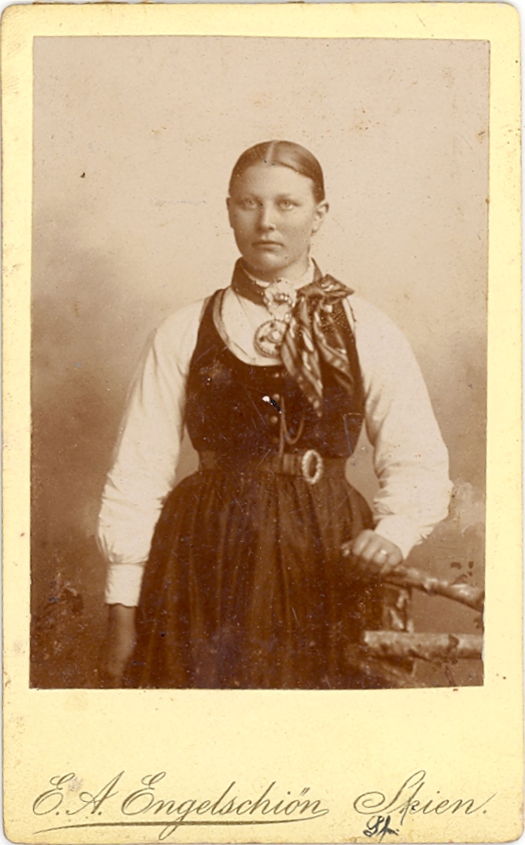 Foto av yngre kvinne i variant av seljorddrakt i atelier.
Kvinna er ukjent.
