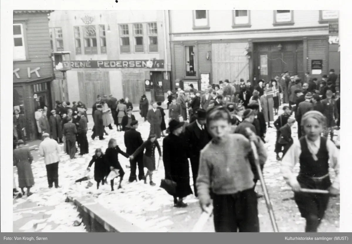 Voksne og barn feirer freden i  Stavanger sentrum kvelden 7. mai 1945. Gatene flyter av arkivmateriale fra Nasjonal samlings kontor. 
I bakgrunnen se forretningen Brødrene Pedersen A/S i Breigata.