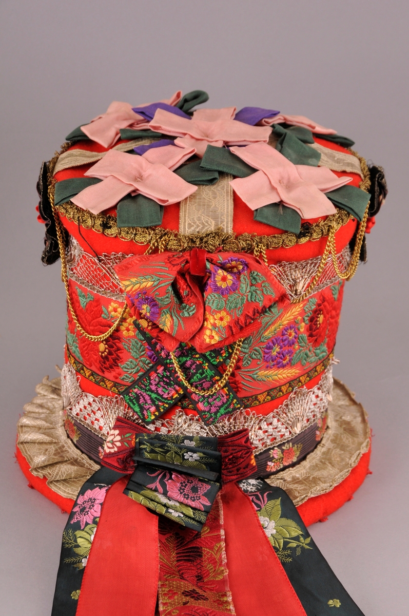 Brudehatt, nylaga. Oppbygd rundt ein filthatt av Zinkes mote AS. Langs bremmens ytterkant er det lagt ei "pølse" av vatt før heile hatten er kledd med høgraudt klede med tydleg lerretsvev. Inni går den raude kleden litt over halvvegs opp i hatten. Der er det  sydd ein løpegang med eit band til å dra saman vidda med, dette for at hatten skal slutte betre rundt hovudet. 
På bremmens overside er ei sylvbore foldelagt og sydd fast langs innerkanten, denne bora er truleg ikkje ny. Vidare rundt hattens pull er det sydd fast eit band med lansert blomemønster, ein sylvknipling, eit smalt band med brodsjert mønster og ein sylvblonde (ulik den som er lenger ned) og til slutt ei avklypt gullblonde/agraman heilt på toppen av pullens kant. Banda er i ein blandingskvalitet.
Over pullens topp (flat del) er ei sylvbore lagt i kryss, over denne er det festa rosettar sydd av grønt, lilla og rosa syntetisk band. Langs kanten av pullen er det sydd fast 3 rosettar, ein midt framme og ein på kvar side. I tillegg er eit "gull"-kjede av uedelt metall sydd fast så det heng i ujamne bogar.
Midt bak på hatten er det øverst sydd fast ei sløyfe av eit lansert bomullsband og ei mindre sløyfe nederst av eit mønstervove band. i tillegg ni lange heng av band, både einsfarga og mønstervove.