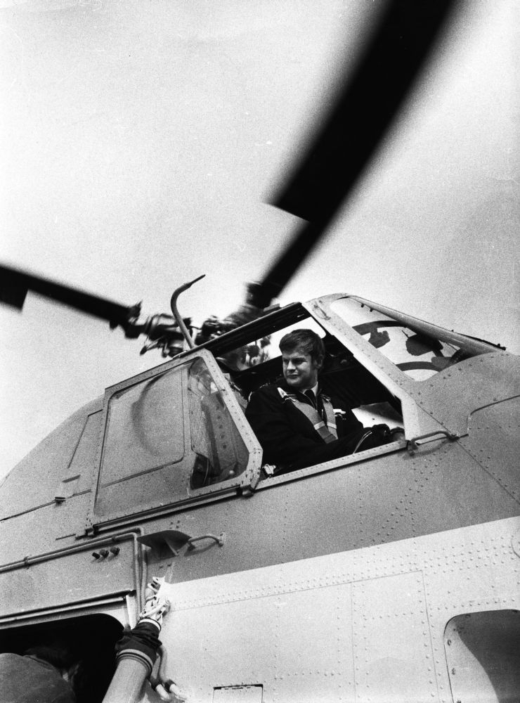 Lufthavn/Flyplass. En person, flyger/pilot, ser ut gjennom en åpen siderute fra cockpit i et helikopter.