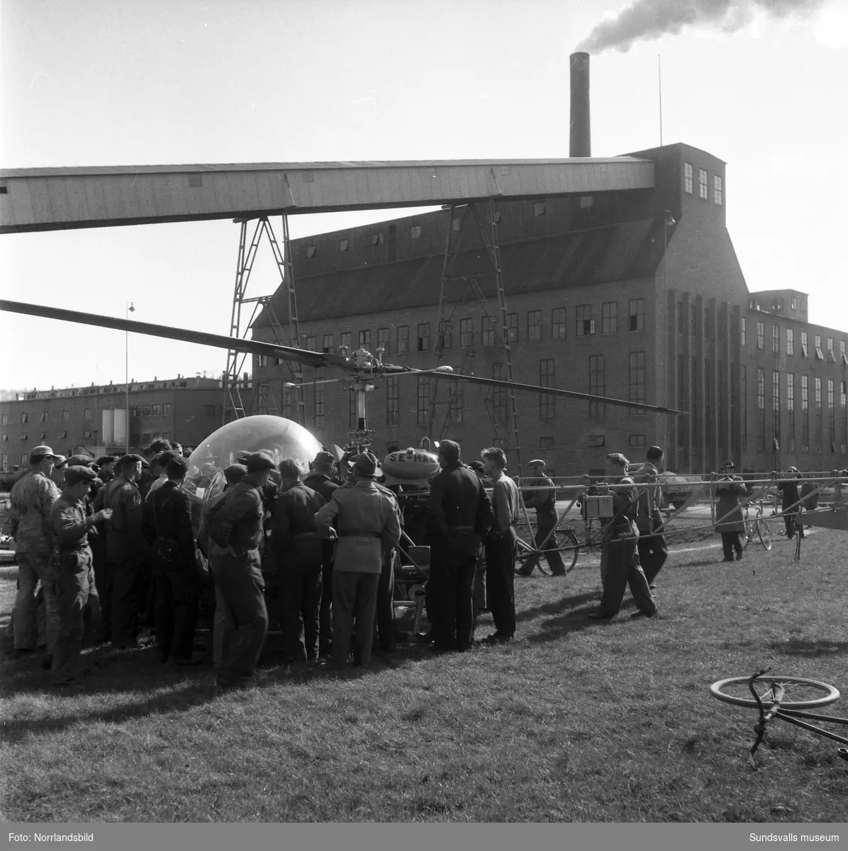 Helikopteruppstigning vid Östrandsfabriken. En helikopter flyger nära det rykande skorstenarna och på marken är intresset stort.