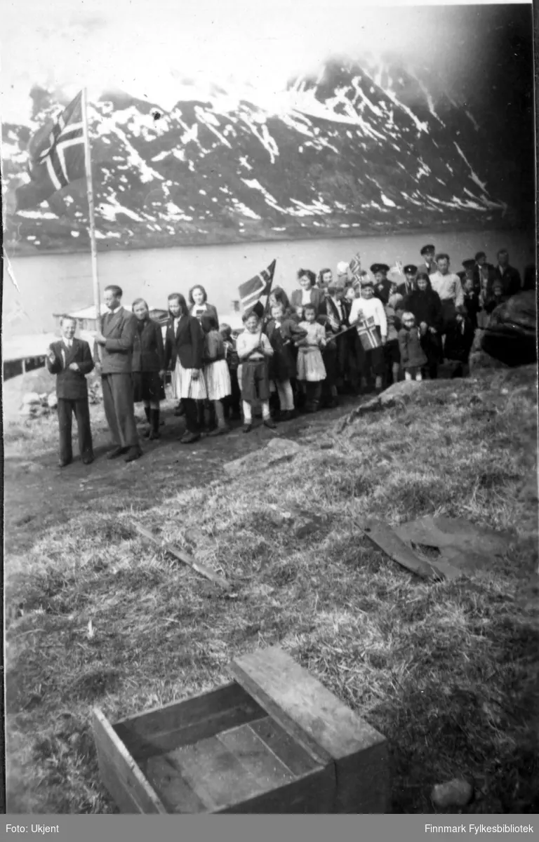 17.mai feiring i Nuvsvåg etter frigjøringen. Fotografiet tatt på slutten av 1940-tallet. Barn går i 17.mai tog med norskeflagg. Barna er kledd i skjørt, luer, bukser, gensere,jakker og sko. En mann i dress holder på et stort flagg helt framst i toget.