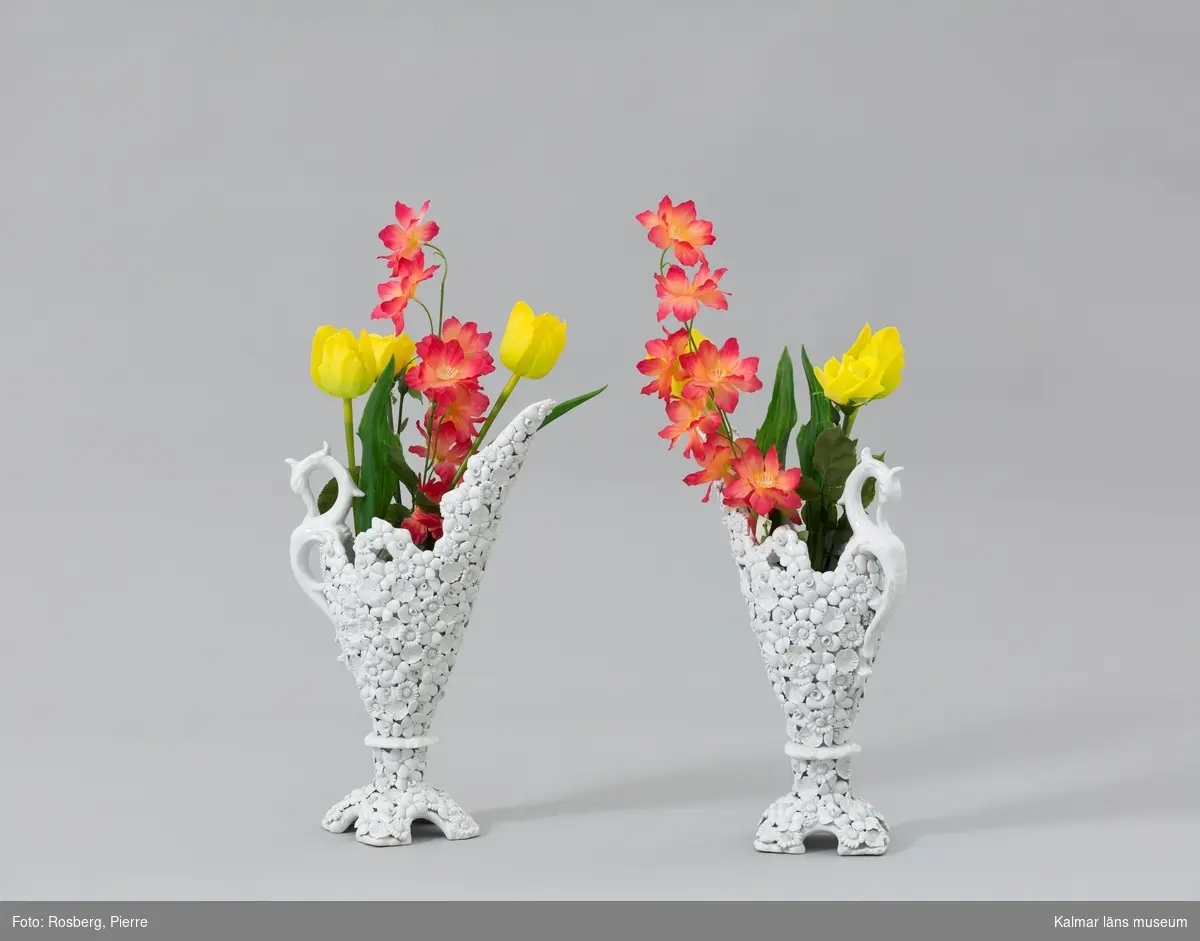 KLM 45619:145:1:1-2. Vas, med plastblomma. Vas (:1) av vitglaserad keramik med ett handtag formad som en sjöhäst. Vasen är formad som en kanna med pip och fot, utsidan är smyckad med blommor och blad. På undersidan en stämpel: ITALY. Till vasen hör ett knippe plastblommor (:2) med bl.a. tulpaner och rosor i gult, rött och grönt.