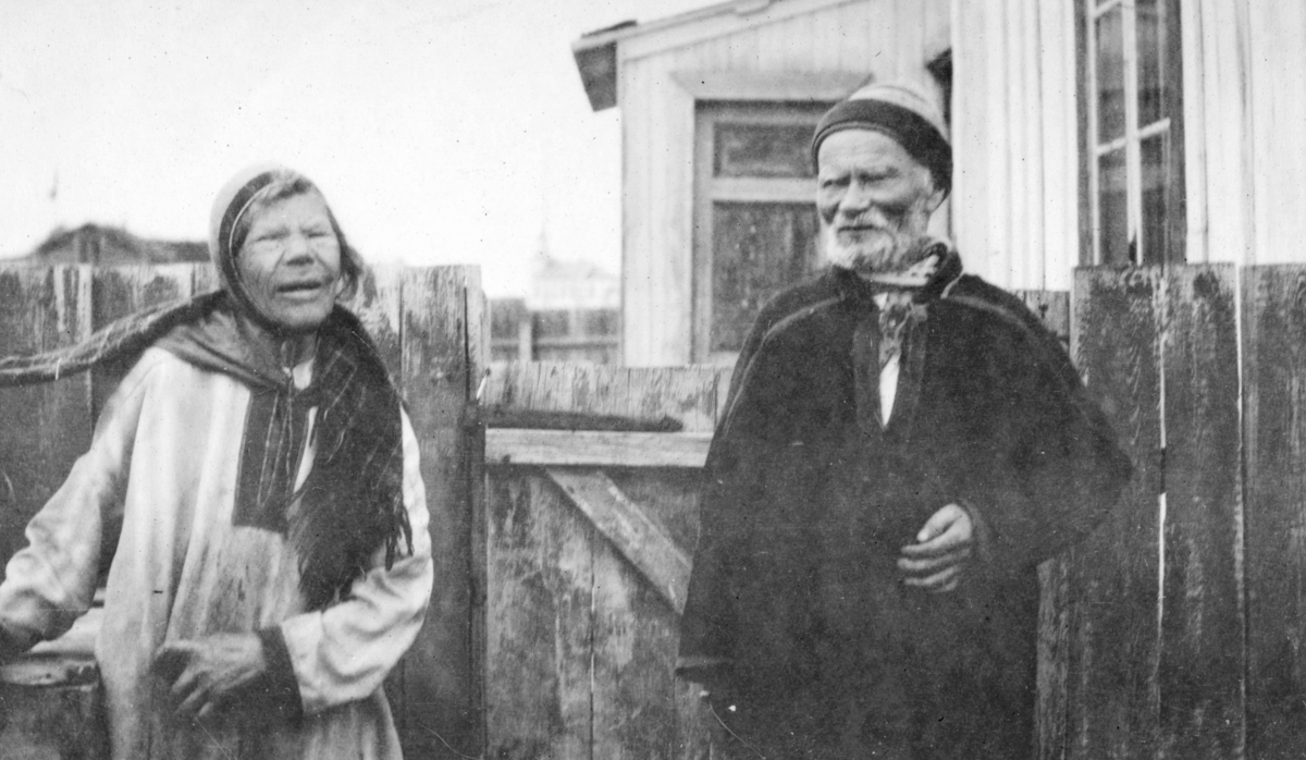 "Akkedas mellem mand og kjærring paa Lebesby gamlehjem". Et eldre ektepar utenfor gamlehjemmet i Lebesby. De er begge kledt i kofter og luer. Kvinnen har et vevd samisk sjal.