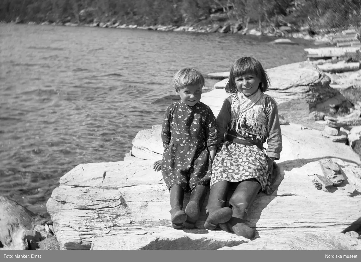 Vid Mellersta vistet i Ritsemjokk. Två flickor, Knut Sjaunas döttrar i blommiga klänningar, sitter på en sten vid vattnet. Sjaunas var den enda familjen i Ritsemjokk som inte bar samisk dräkt till vardags.