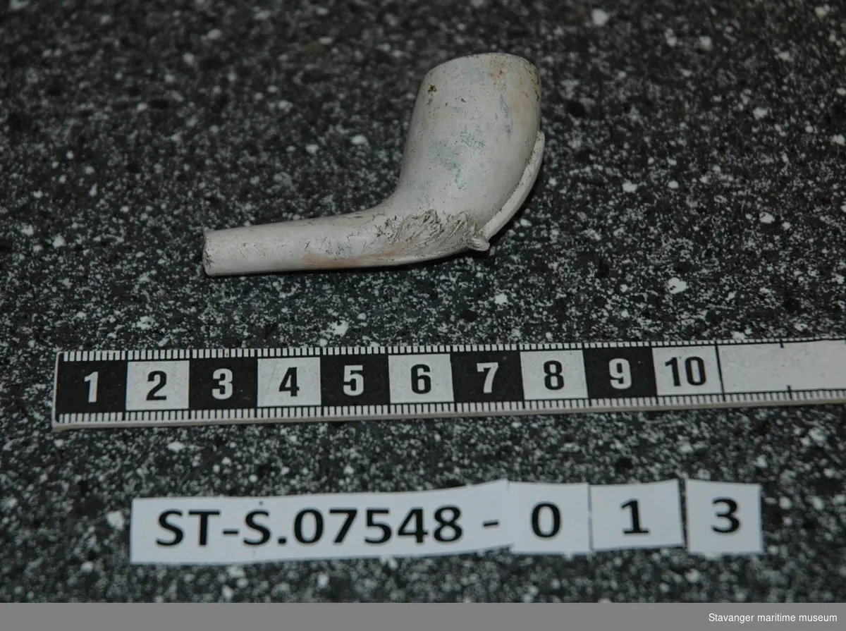 Krittpipehode med fragment av stilk. Funnet i sjakt 2. Dekorert på undersiden med løvblad-lignende utskjæring. Funnet på ca. 30cm dybde.
