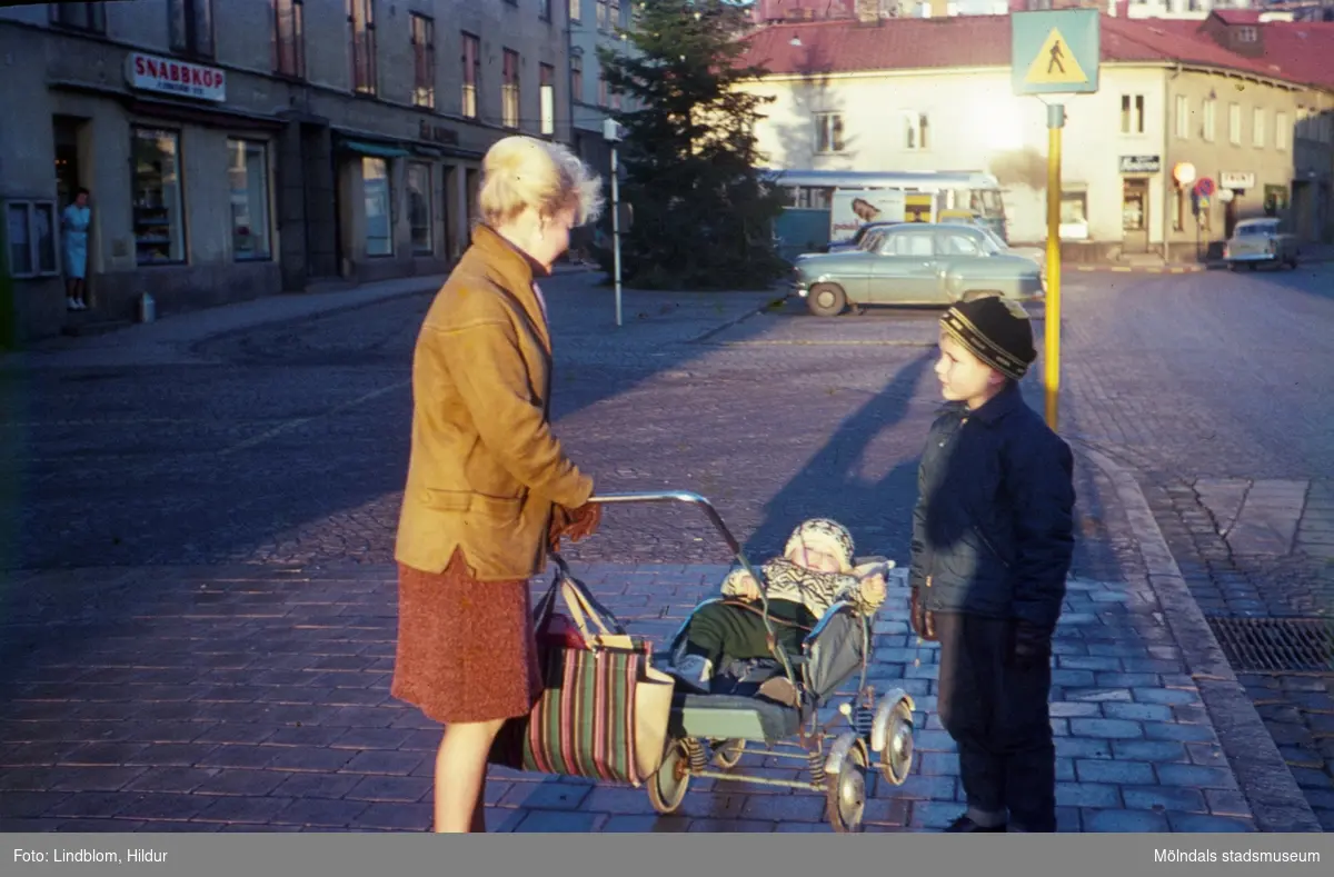 En kvinna med två barn, varav det ena ligger i en barnvagn, står på Gamla Torget i Mölndal, 1960-tal. I bakgrunden till vänster ses huset Kvarnbygatan 43, tidigare Mölndals stadshus, sedermera musikskola. Till höger ses Kvarnbygatan 45.

För mer information om bilden se under tilläggsinformation.