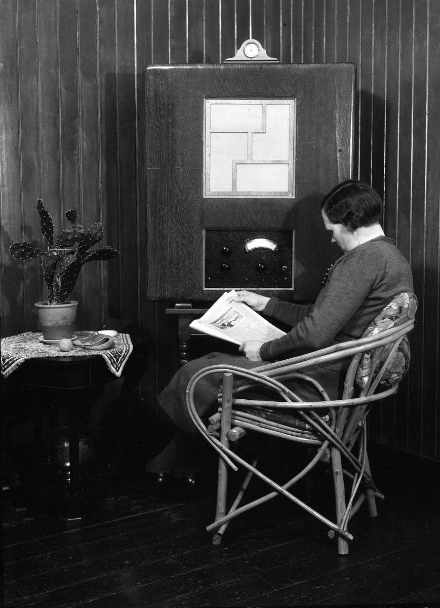 Karine Røisli leser avis foran radioen. Avisen er merket "Torsdag 9.april", noe som tilsier at årstallet er 1936.