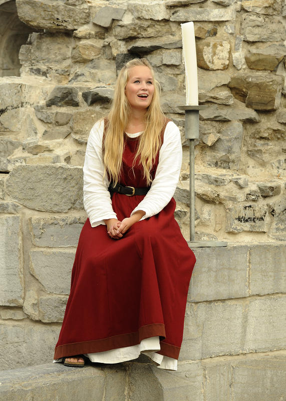 Syngende guide i middelalderdrakt (rød ull overkjole uten erm og hvit linkjole med lang erm under) sitter i domkirkeruinen og synger.
