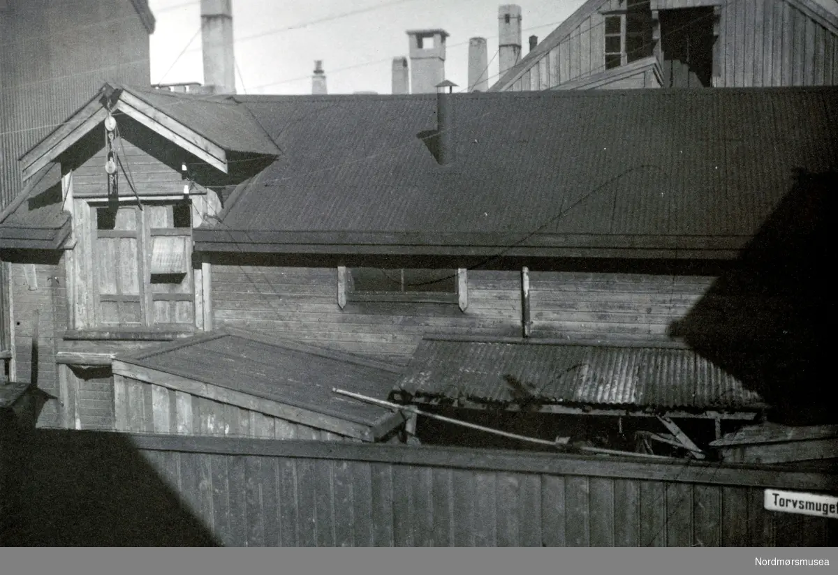 Foto fra Torvsmuget, trolig i Kristiansund, hvor vi ser fra tak og skorsteinspiper, samt en vegg som bærer gatetskiltet. Fra Nordmøre museums fotosamlinger. Reg: EFR
