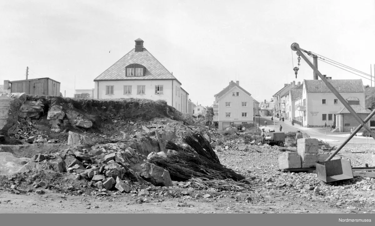 Siste rest av fjellhaugen der Arbeiderforeningen og Rådstua stod. I bakgrunnen ser vi rester av en tysk bunker som ble bygget under krigen 1940-45, og over denne bunkeren hadde da Arbeiderforeningen stått før bybrannen i de sene aprildagene 1940. Bak denne igjen ser vi det nye bygg for folkebiblioteket. I bakgrunnen ser vi Indremisjonsbygget og oppover Marstrands gate. Badet (seinere Nor-Pro) til høyre, revet (først parkering for ny-banken, pr 2007 boligblokk). Her holdt lastebilsentralen til i 1960-åra, pr 2007 Hydro bensinstasjon, og drosjene er fortsatt pr 2007 foran Misjonshuset. 
Inngangen støpt igjen i januar 2017 i forb. med butikk i Tinghuset. ( Regelbau 633 for M19 bombekaster) (info fra Bjørn Ove Angvik - 30.01.2017)
 

Nordmøre Museums fotosamling