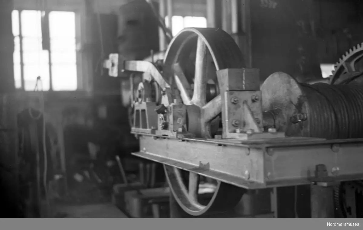 Bildet viser heismaskineriet på den nye bukk-krana klinkbygget i stål ca. 1945 og revet ca. 1970 og sto på kjelkrankaia på Storviks Mek. Verksted. Bukk-krana ble kallt ";kjelkrana"; fordi den ble brukt til tungløftene med blant annet dampkjelene og dampmaskinene i nybyggene og ombyggingene. Bildet er tatt i nedre del av maskinverkstedet før montasje og man ser bremsearmen og bremsetrommelen og wiretrommelen. I bakgrunnen ses en av de sylindriske vedovnene, med luftehuller, i maskiverkstedet. Kjelkrana ble også brukt til å løfte ut kjelene og dampmaskinene på sidetrålerne ved skifte til motor. Kranen hadde en mangeskåret løftewire og en løfteevne på 25 tonn og var den kraftigste i Kristiansundsområdet. Dette er samme heisverket som FAKf-100296.171645 og FAKf-100296-186086. Tekst av Peter Storvik. Fra Nordmøre museums fotosamlinger.
