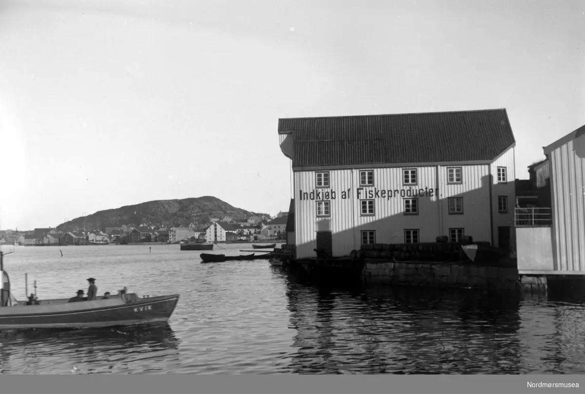 Et bilde av sundbåten Kvik som er på vei til kai på Innlandet, i Kristiansund. ";Indkjøb af Fiskeproducter"; står det på bryggeveggen (Kjønnøybrygga, pr 2008 leid av Rica hotel). Gamle Tollbua til høyre. Nordmøre Museums fotosamlinger
