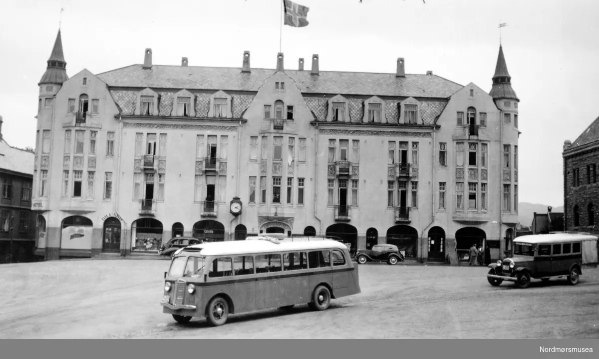 Dette bildet stammer nok tidligst fra 1935. Bussen er sikkert T-4232, en International, 1935-modell, som ble anskaffet av Kristiansund-Oppdal Auto (KOA) for å gå i rute Oppdal-Kristiansund. Se R.Fot.42756 for bedre bilde av bussen. 
Det var først i 1932 at KOA og Surnadal Billag kjørte ruter helt fram til Kristiansund. Det skjedde først da det ble satt inn bilferge Kvisvik-Kristiansund. Først i 1931 ble veien utover fra Sunndalsøra og Tingvoll åpnet helt fram til Kvisvik. Bussen bak til høyre kan også stamme fra KOA. 
Selskapet hadde ruter Kristiansund-Oppdal i korrespondanse med nattog til og fra Oslo. Det var et tilbud for passasjerer og post. (Info: 
Sveinung Berild). - To busser på Torget på Kirkelandet i Kristiansund. I bakgrunnen ser vi Grand Hotell (nummer to). Fra Nordmøre museums fotosamlinger.