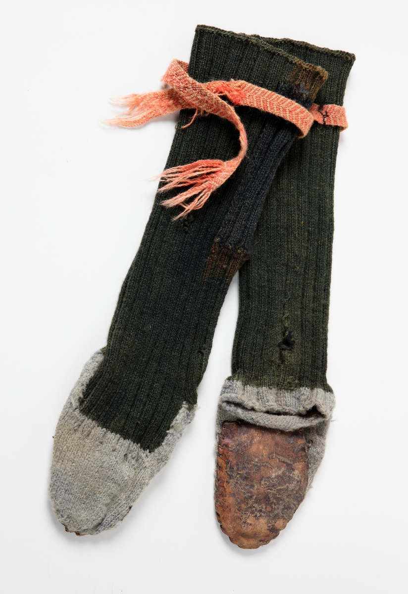 Svarte raggsokker i ull med såler av lær. Fot av grått ullgern, uten hæl.
Sokkebånd flettet av rødt og gult ullgarn, fiskebeinmønster.