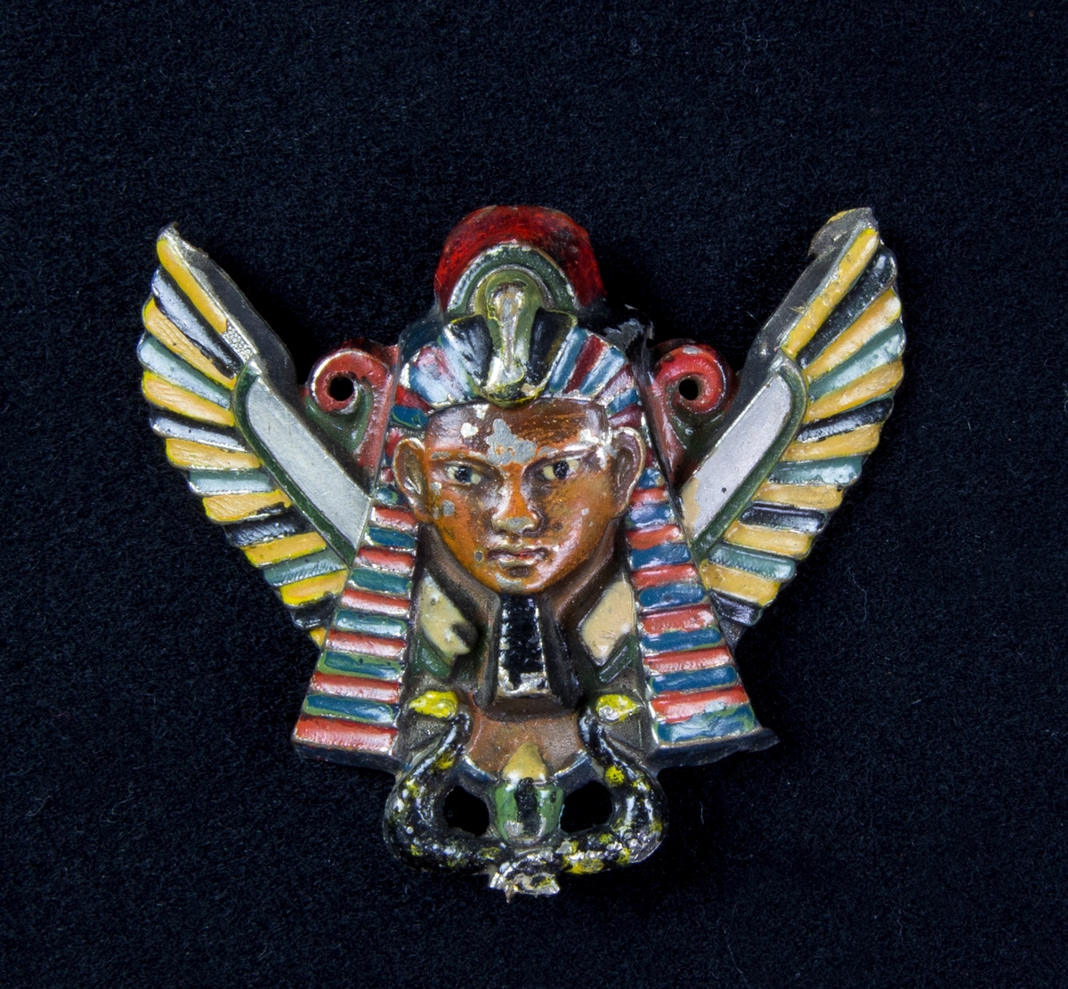 Beslag av vitmetall i egyptisk stil i form av en farao, fågel med utbredda vingar och två ormar i rött, grönt och gult. Två hål att fästa på klädesplagg.