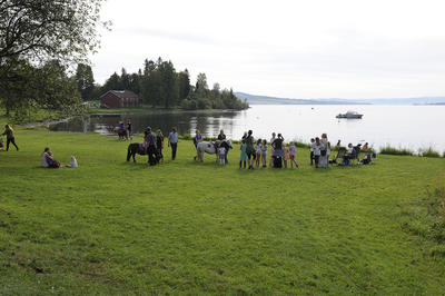 Barn og ungdom som driver med aktivitet på grønn gresslette ved vannet, museumshus i bakgrunnen.. Foto/Photo