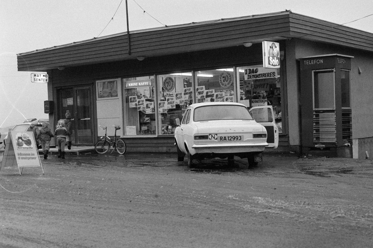 Parkert bil og barn utenfor butikk på Berg. Telefonkiosk til høyre.