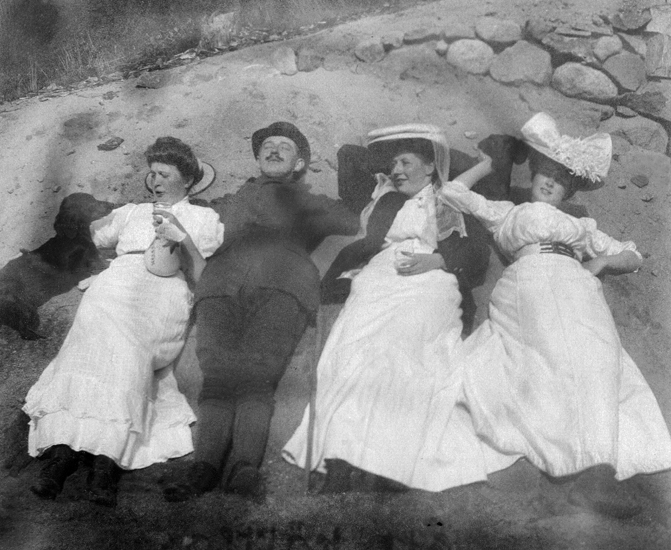 Fyra personer (tre kvinnor och en man), ligger i solen på en sandvall, tillsammans med två släthåriga taxar.