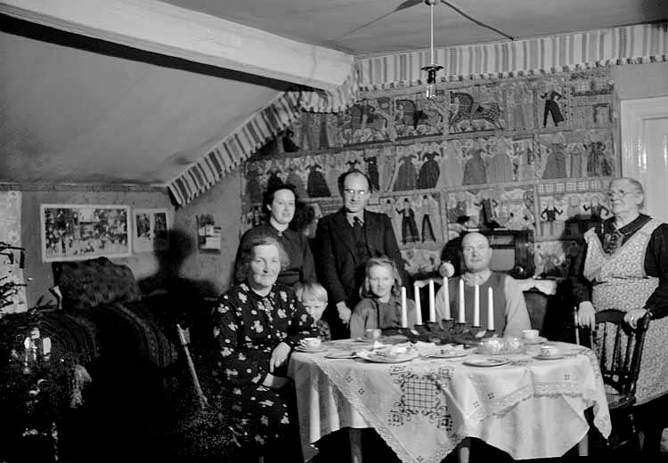 En familj som samlats vid julbordet, de dricker kaffe.
På väggen hänger en stor bonad med olika bilder. Till vänster skymtar en julgran.