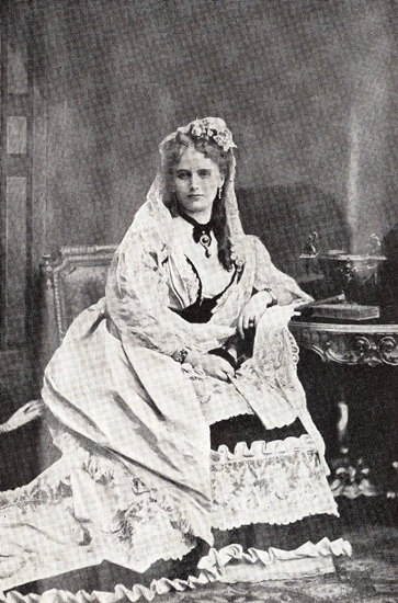 Porträttfoto av Christina Nilsson, ca 1875, Hon sitter vid ett bord med en klänning på sig samt bär någon form av slöja med blommor.