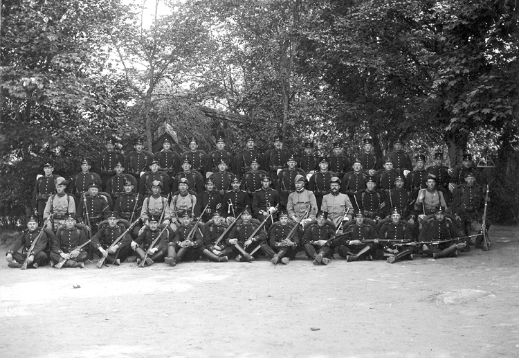 Ett kompani har ställt upp sig för fotografering på en grusplan framför de berömda hyddorna, som skymtar i bakgrunden. Officerarna sitter på1:a raden, i mitten.En del har den äldre uniformen, några har m/10 med trekantiga hattar m.m.Kapt. Westin - Erik Westin (1871-1914), kapten m.m. v. Kronobergs regemente.Sune Nordenskjöld - (1881-1957), löjtnant v. Kronobergs regemente.Senare överstelöjtnant, Kristianstad.Källa: Svensk Rikskalender 1908, s. 173. Svensk Adelskalender 1904,1936, 1949. Sveriges Dödbok 1947-2003.