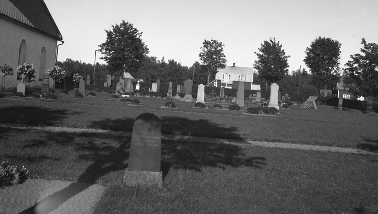 Foto på kyrkogård med gravstenar. Jämte finns en kyrka och  längre bort står det bostadshus.