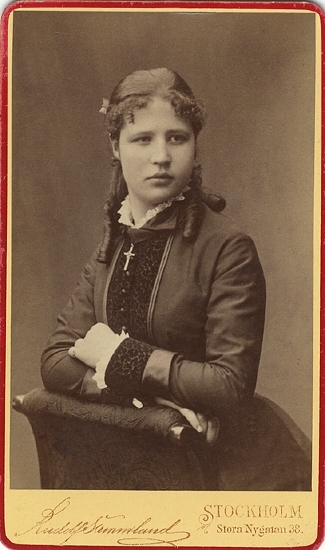 Porträtt (midjebild, halvprofil) av en ung, okänd kvinna i mörk klänning med sammetsbrokad och vit kråskrage. 
Vid kragen syns en kedja med ett kors. 
Hon stöder armarna mot ryggstödet på en stoppad stol.