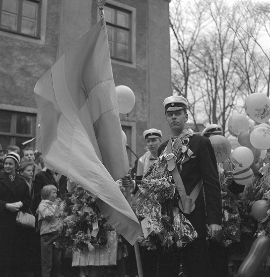 Studenterna första dagen, 1959. 
En fanbärare m.fl. vid Esaias Tegnérs staty. I bakgrunden skymtar Karolinerhuset.