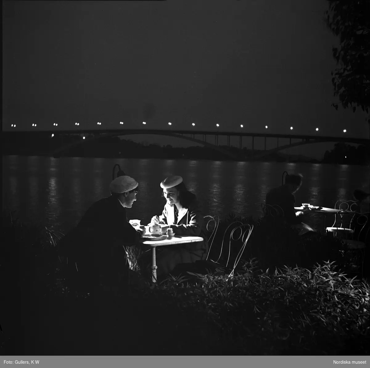 Norr Mälarstrand, Stockholm. Två kvinnor sitter vid ett bord på en uteservering i kvällsbelysning. Riddarfjärden och Västerbron i bakgrunden.