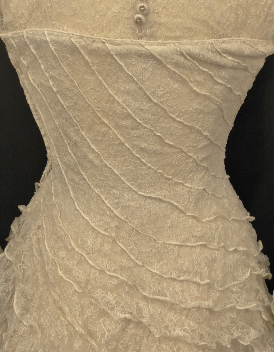 Helklipt, figursydd. Ermer og bærestykket klipt i ett. Rund hals bak med 22,5 cm lang splitt og 14 knapper med sydde trådhemper. Foran er halsutringingen rett nedover 18,5 cm fra skulderbredden ved halsen og går ned under bryststykket på kjolen. Fra bærestykket (som er foret med bobinett) er kjolen foret med tykk, blank silke. Utenpå er påsydd bobinettkapper som går på skrå fra venstre og ned mot høgre. Størsteparten av overdelen og hele skjørtet er blonden sydd på bobinettstoff uten mønster. Underskjørtet er da løst og har ei snor innsydd i folden nede for at kjolen skal føre seg penere. Glidelås i siden, hekter og sydde hemper på overdelen. Erme: Skinkeerme med 12,5 cm lang splitt ved håndleddet og 7 knapper, sydde knappehemper. 6 små innsydde legg på underarmen for bue av ermet. Skjørtet er utsvingt og har ein vidde nede på ca. 668 cm. Vidden på underskjørtet 470 cm. Kjolen er 19 cm lengre bak enn foran.