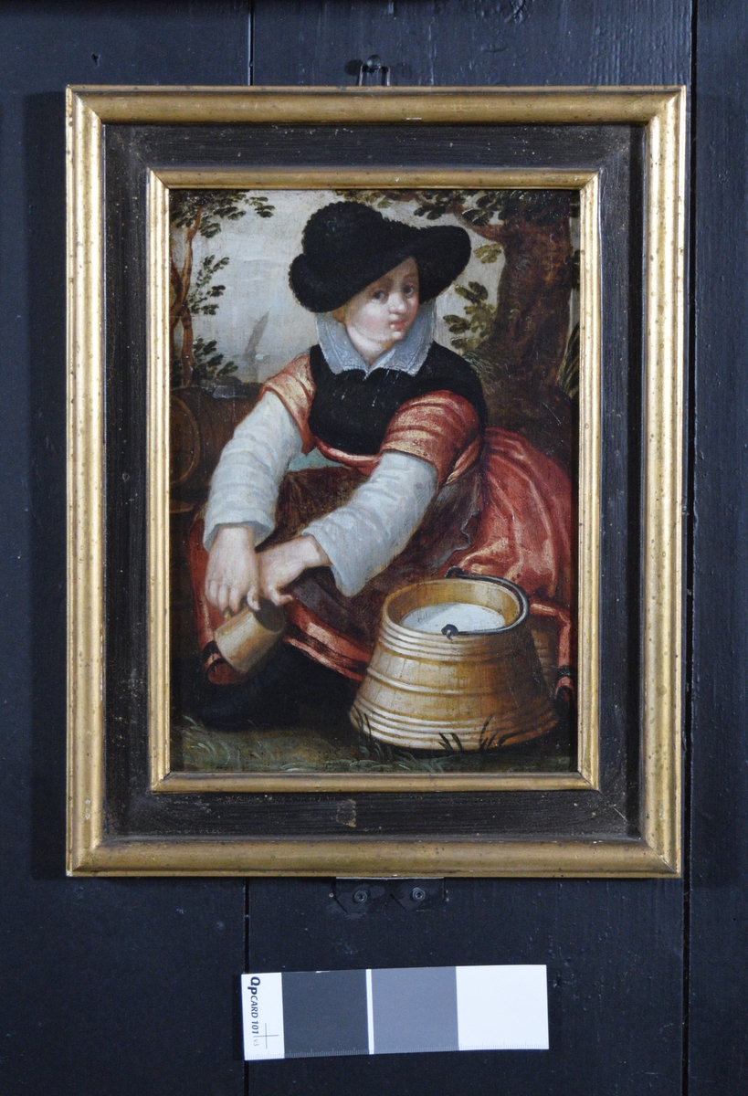 Bildet viser en stillesittende bondepike i rød kjole og sort hatt. I hendene som henger over knærne holder hun et krus. Foran henne står et lagget spann med melk i. I bakgrunnen ser man en tønne, trær og lengre borte en båt eller pram under seil.