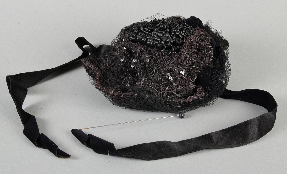 Damhatt av babymodell av svart strå med svart flor, påsydda svarta paljetter, svarta sammetsrosetter och band. Knytband av svart sammet. Fodrad med svart siden.