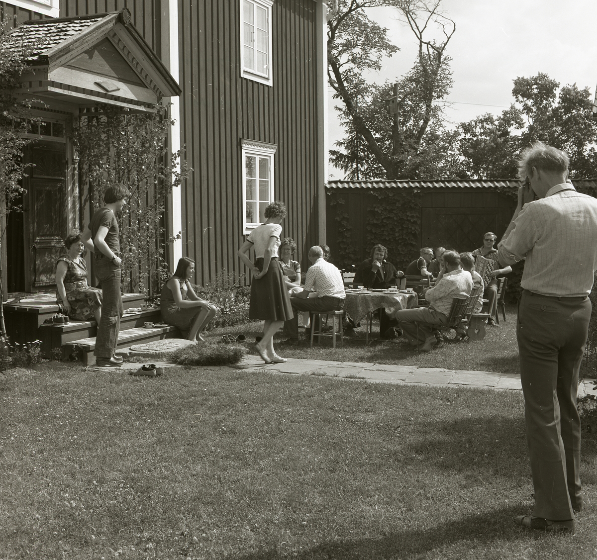 I trädgårdsmöblerna på gården Sunnanåker är ett sällskap bänkade för en kaffetår midsommarafton 1978. Intill husväggen har gruppen funnit en solig plats för avkoppling och underhållning då en av männen spelar dragspel.