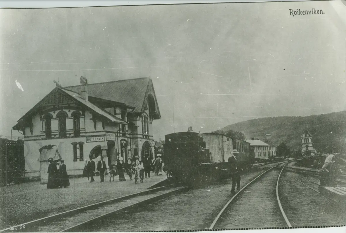 Røykenvik jernbanestasjon med tog og en rekke personer på perrongen, aviskiosk. Vanntårn i bakgrunnen. 