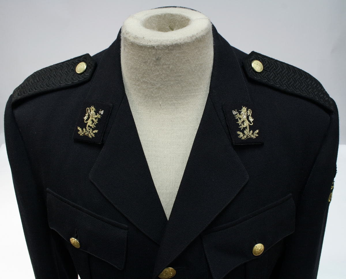 Enkeltkneppet jakke med fire riksløveknapper, fire ytterlommer, broderte riksløver på kragespeil, politimerke på ermet, og sorte skulderklaffer.