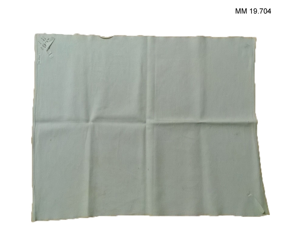 Vävd vit handduk i så kallad gåsöga mönster. Maskinfållad i kortändarna. I ena hörnet stämplad: 1.11.1937 och påsydd hank av vitt bomullsband. I hörnet diagonalt mitt över, även där en hank av bomullsband.
Ett par svaga stämplar: Tre Kronor i handdukens nedre del.