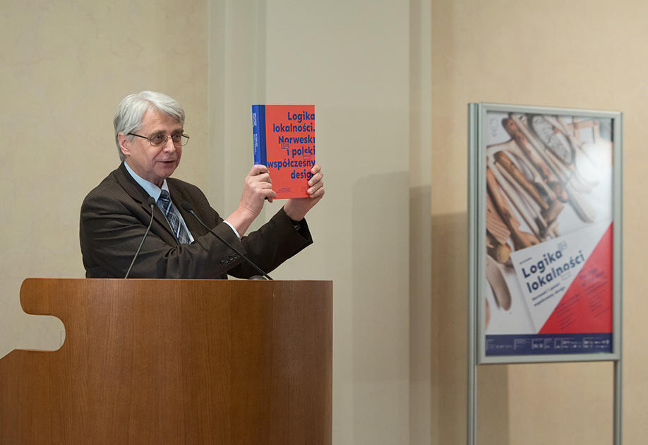 Prof. Dr. Jacek Purchla, direktør ved ICC, står bak talerstolen og holder opp utstillingskatalogen på åpningen av Logika Lokalnosci, 16. desember 2016. Plakaten for utstillingen sees i bakgrunnen. (Foto/Photo)