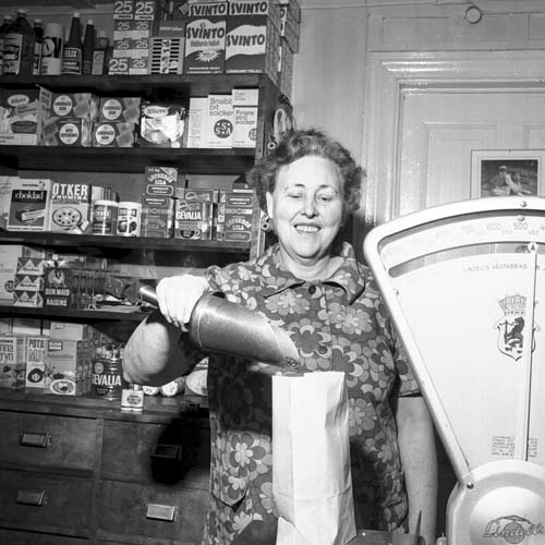 Astrid Lindblom står i sin butik 1970 och öser upp troligen kaffebönor i en papperspåse, vågen visar på ett halvkilo. Bakom henne i vägghyllorna står olika förpackningar stålull, kaffe, potatismjöl och bitsocker