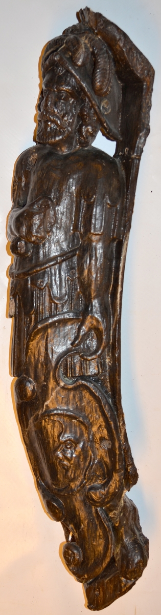 Krigare iklädd romersk rustning samt plymagerad hjälm. Mannen har långt hår och skägg. I höger hand håller mannen ett draget svärd snett över bröstet och upp mot vänster axel. Svärdsklingan är snidad separat. Vänster hand fattar om en stor, profilerad sköld. Skölden är dekorerad med en i relief skuren halv mångubbe. Skölden vilar mot vänster ben. Mannens huvud och bål är kraftigt vridna mot vänster medan underkropp och ben är placerade i en face. Höger ben är placerat framför vänster ben som till större delen är dold bakom skölden. Benen är stövelklädda. Utrymmet för fötterna är minimalt.

Figuren är placerad snett i förhållande till bakstycket. Skulpturens fotända är, sedd framifrån, i det närmaste trekantig. Skulpturens baksida visar tydliga huggspår och har ett snett urtag upptill som lämnar huvudändan nedsänkt. En markant urgröpning med rektangulärt hål kan iakttas på detta parti.

Skulpturen och bakstycket är snidat i ett stycke. Armarna är snidade separat och sitter fästade vid bålen med dymlingar. Bakstycket är profilsvängt och baksidan är slät. Nedtill är bakstycket framdraget så att det bildas en kraftig sockel på vilken figuren står. Sockeln är ornerad på sidorna. Bakstyckets övre del fortsätter snett uppåt och bildar en bred platta ovanför krigarens huvud.
Skulpturen är relativt välbevarad. Skulpturens baksida och fotända är delvist skadade.

Text in English: Warrior clad in Roman armour, with long hair and a beard. A simple plumed helmet adorns his head. The man draws his sword with his right hand across his chest and up towards his left shoulder. The blade of the sword was carved separately. The left hand grasps a large, profiled shield, decorated in relief with a profile of half the face of the moon. The shield rests against the left leg. The man''s head and body are twisted considerably to the left. The shield conceals most of the body.

The figure is placed at an angle in relation to the back-board. The foot end of the sculpture is practically triangular as seen from the front. the rear side of the sculpture has a slanted groove near the top that leaves the head end lower, indicating that the sculpture is attached partly to the outside planking.

The sculpture is carved from a single piece of wood, except for the arms which are carved separately and attached to the thorax with dowels. The figure is placed in front of a profiled back-board, with a smooth reverse side, which is drawn forward at the bottom to form a robust base upon which the figure stands. The base is simply ornamented on the sides. At the top the back-board continues above the heads to form a wide plate-like section over the warrior''s head.
The sculpture is rather well preserved.  Skulpturens baksida och fotända är delvist skadade.